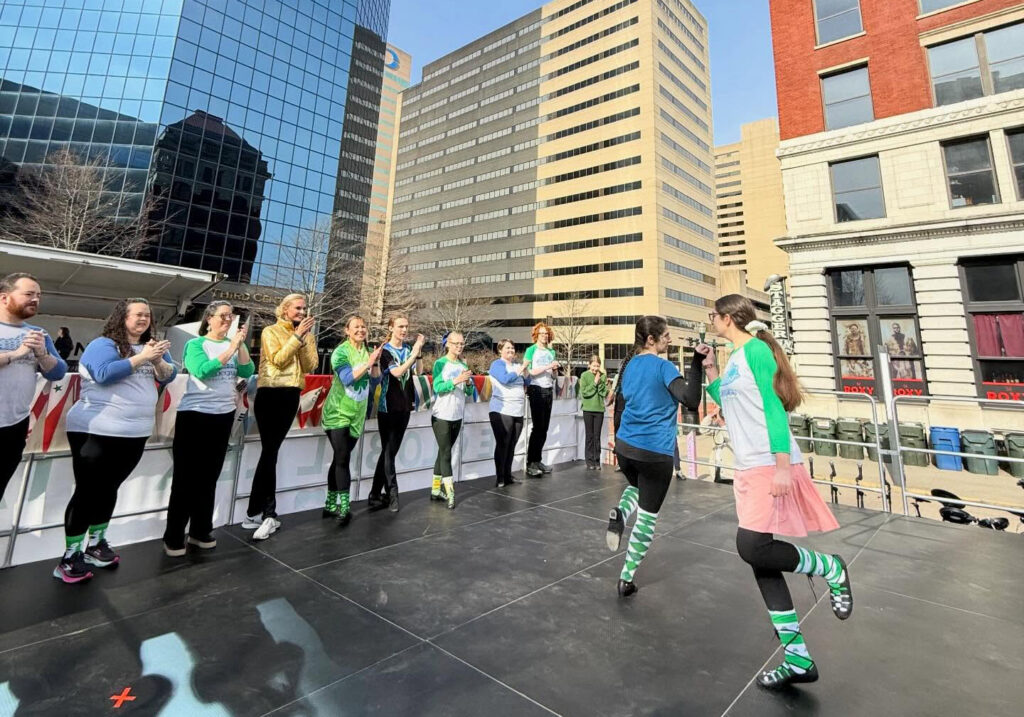 Two dancers from our Irish dance classes in Lexington performing a routine in soft shoes on an outdoor stage in downtown Lexington while fellow Bluegrass Ceili Academy dancers clap along.
