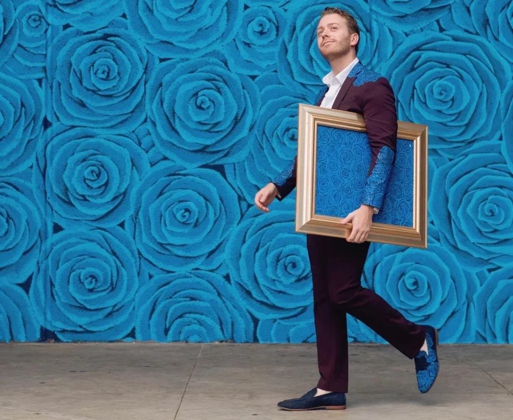 Artist Wylie Caudill carrying a gold framed painting in front of his iconic cerulean blue rose mural wall in Lexington, Kentucky